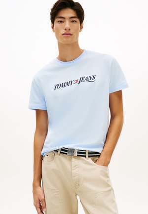 Νεαρός άνδρας που φοράει ανοιχτό μπλε μπλούζα Tommy Jeans και μπεζ παντελόνι με ριγέ ζώνη, στέκεται με το ένα χέρι στην τσέπη.