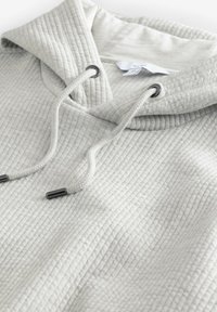 Sudadera con capucha de color gris claro, fabricada con un tejido texturizado y acanalado. Cuenta con cordones con puntas de metal y un bolsillo frontal.