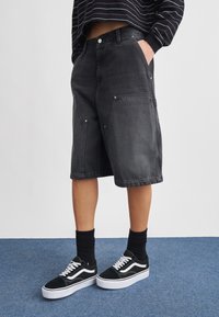 Άτομο που φοράει μαύρα τζιν σορτς μέχρι το γόνατο, μαύρες κάλτσες και μαύρα-λευκά αθλητικά παπούτσια, στέκεται σε μπλε χαλί μπροστά σε λευκό τοίχο.