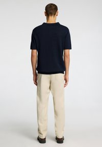 Navy polo shirt with short sleeves, textured fabric, and a classic collar, paired with beige trousers and dark shoes, viewed from the back.