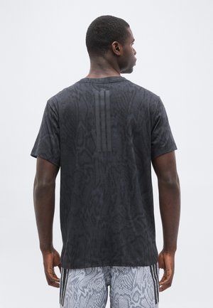 Mand vendt bagud iført en sort mønstret T-shirt med tre lodrette striber på den øverste del af ryggen og lyse mønstrede shorts med lodrette striber.