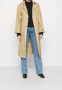 Persona che indossa un lungo trench coat beige sopra un maglione a collo alto nero, jeans blu e stivaletti neri, in piedi contro uno sfondo bianco.