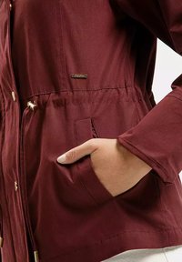 Burgundowa lekkka kurtka z możliwością regulacji w talii, z złotymi elementami, kieszenią z przodu oraz długimi rękawami dla luźniejszego fasonu.