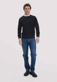 Schwarzer Strickpullover mit Rundhalsausschnitt, blaue Jeans und schwarze Stiefel. Das Model steht vor einem hellen Hintergrund und präsentiert das Outfit.