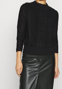 Femme portant un pull en tricot noir à manches longues avec un devant à volants et de petits boutons, assorti à une jupe en cuir noire.