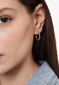 Orecchini a cerchio in argento con un design geometrico, caratterizzati da piccole pietre preziose trasparenti lungo il bordo, indossati all'orecchio sinistro.