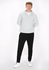 Hellgrauer Pullover mit einem halben Reißverschluss am Kragen, kombiniert mit schwarzen Jogginghosen und weißen Sneakers. Glattes Material, lockere Passform, einfaches Design.