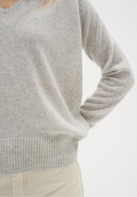 Personne portant un pull en V gris clair avec ourlet côtelé et détails de couture latérale, associée à un pantalon beige clair, main sur la hanche.
