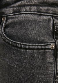 Vaqueros de mezclilla en gris oscuro con un bolsillo delantero, costuras visibles y un detalle de botón de metal. La tela muestra una textura y un patrón sutil.