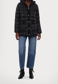 Chaqueta acolchada negra con capucha, que presenta costuras horizontales, bolsillos frontales, cierre de cremallera y herrajes plateados. Combinada con un par de jeans azules y botas negras.