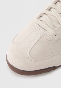 Μπεζ σουέντ αθλητικό παπούτσι με υφασμένο επάνω μέρος, στρογγυλεμένη μύτη και ανοιχτό μπεζ κορδόνια. Η σόλα είναι καφέ καουτσούκ με λείο φινίρισμα.