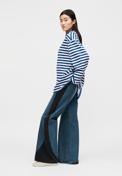 Νέα γυναίκα με μακρυμάνικη μπλούζα με μπλε και λευκές ρίγες, φαρδιά μπλε τζιν με μαύρα πάνελ και λευκά αθλητικά παπούτσια, στέκεται πλαγίως.