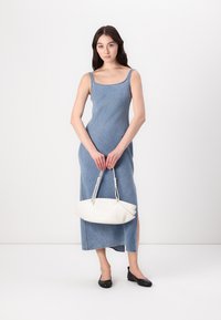 Denim maxi šaty s tenkými ramienkami, priamou siluetou a bočným rozparkom. Model drží bielu zakrivenú kabelku a má na sebe čierne ploché topánky.