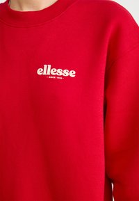 Piros pulóver, kerek nyakkal, pamutból készült, kis fehér "ellesse" logóval és "1959 óta" felirattal a mellrészén.