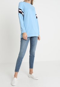 Donna che indossa una felpa a maniche lunghe di colore azzurro chiaro con strisce nere, bianche e borgogna sulle maniche, jeans blu aderenti e scarpe bianche senza lacci.