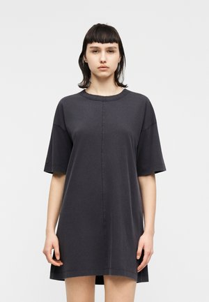 Ung kvinde med mørkt, glat hår iført en løs, kortærmet, mørkegrå kjole, stående foran en ensfarvet baggrund.