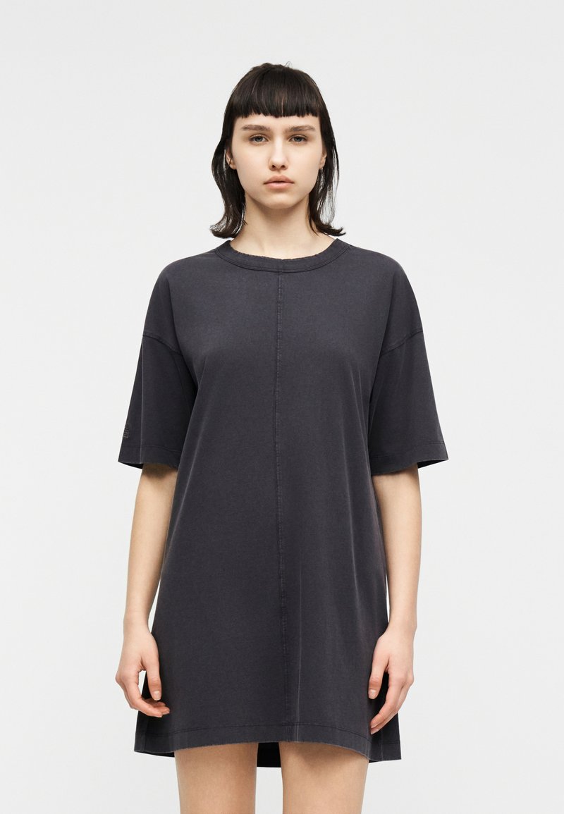 Νεαρή γυναίκα με σκούρα ίσια μαλλιά, φορώντας ένα φαρδύ, κοντομάνικο, σκούρο γκρι φόρεμα, στέκεται μπροστά σε μια απλή φόντο.