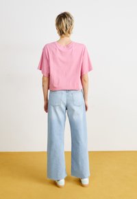 Pink short-sleeve T-shirt, loose cut, paired with light blue wide-leg jeans. Model wears mint-colored shoes. Neutral background with yellow floor.