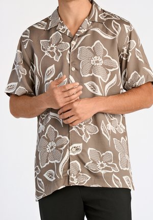 Homme portant une chemise beige à manches courtes avec un grand imprimé floral blanc, mains jointes devant, associé à un pantalon noir, sur un fond uni.
