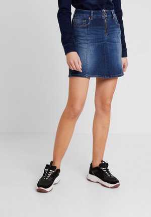Femme portant un chemisier bleu foncé à manches longues, une mini-jupe en denim bleu avec fermeture éclair frontale, et des baskets noires et blanches, debout devant un fond uni.