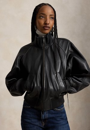 Jeune femme aux longues tresses portant de grandes boucles d'oreilles créoles, une veste en cuir noire zippée, un col roulé blanc et un jean bleu, les mains dans les poches.