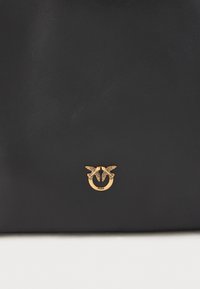 Melna ādas rokassoma ar teksturētu virsmu un zelta toņa logo detaļu, uz kuras ir divi putni, kuriem apkārt ir gredzens.