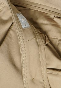 Beige tyg med sömdetaljer, dragkedjestängning och en liten knapp. Har en slät yta och en ren finish.