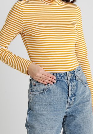 Frau trägt einen senfgelb-weiß gestreiften langärmeligen Rollkragenpullover, in hoch taillierte blaue Jeans gesteckt, Hand auf der Hüfte ruhend.