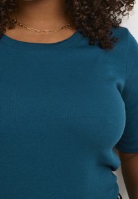 Teal ribbet strik T-shirt med korte ærmer og rund halsudskæring. Glat tekstur med subtil stræk. Guld kæde halskæde som accent.