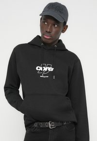 Νέος άνδρας που φορεί μαύρο φούτερ με κουκούλα και λευκό λογότυπο "core" και σκούρο γκρι καπέλο, κοιτάζοντας προς το πλάι, με τα χέρια στην τσέπη του φουτερού.