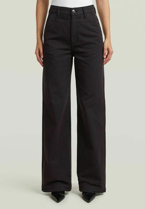 Personne portant un pantalon noir taille haute à larges jambes, avec fermeture par bouton et zip, associé à des chaussures noires pointues et un haut blanc.