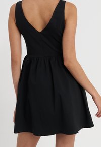 Robe noire avec un corsage ajusté et une jupe évasée. Caractérisée par un dos en V profond et des bretelles larges. Confectionnée dans un tissu lisse avec une texture minimale.