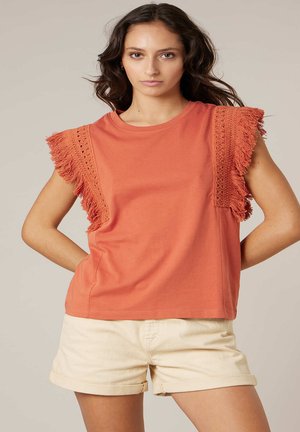 Femme portant un haut orange rouille avec des manches en crochet et à franges, et un short beige retroussé, debout les mains sur les hanches.