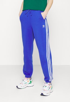 Osoba ubrana w niebieskie spodnie dresowe Adidas z białymi paskami oraz białe trampki z kolorowymi akcentami, stoi z rękami w kieszeniach.