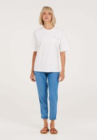 Weißes Baumwoll-T-Shirt mit kurzen Ärmeln, kombiniert mit hellblauen Jeans. Das Model trägt braune Sandalen. Einfaches Design mit minimalen Details.