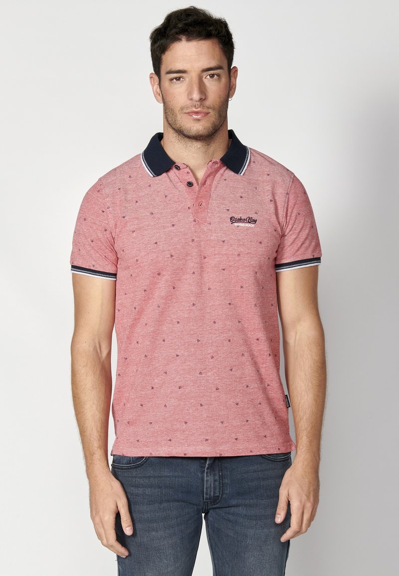 Röd kortärmad polo med marinblå krage, som har ett mönster av små blå trianglar och en logotyp på vänster bröst. Bärs med jeans.