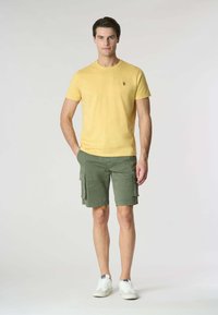 T-shirt giallo a maniche corte con una sottile texture e un piccolo logo, abbinato a pantaloni cargo verdi con numerose tasche. Sneakers bianche.