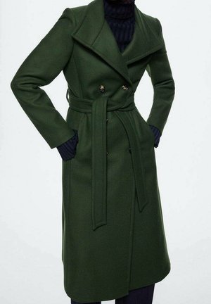 Femme portant un manteau en laine vert foncé ceinturé sur un pull col roulé noir, les mains dans les poches du manteau, debout devant un fond uni.