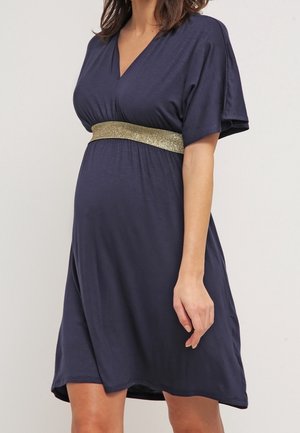 Schwangere Frau trägt ein marineblaues, knielanges Kleid mit V-Ausschnitt und breitem goldenem Taillenbund.