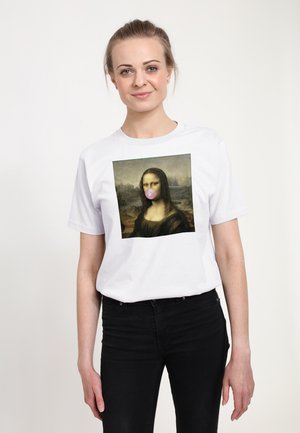Kvinde iført hvid t-shirt med Mona Lisa-kunsttryk, der blæser lyserød tyggegummi boble trykt foran, kombineret med sorte bukser.