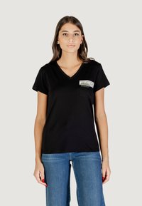 Czarny T-shirt z dekoltem w serek i krótkim rękawem, z małą kieszonk�ą z graficznym wzorem. Gładki materiał o luźnym kroju. Widoczne dżinsy.