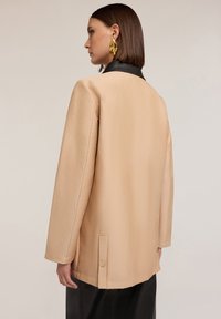 Beige oversize bunda s černým límcem, dlouhými rukávy a knoflíkovým detailem na lemu. Hladká textura, minimalistický design.