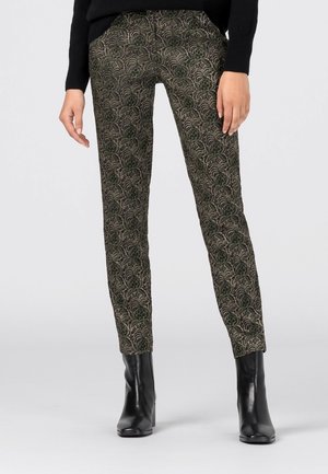 Femme portant un pantalon slim avec un motif complexe de feuilles vertes et noires, associé à des bottines noires à talons et un pull noir.