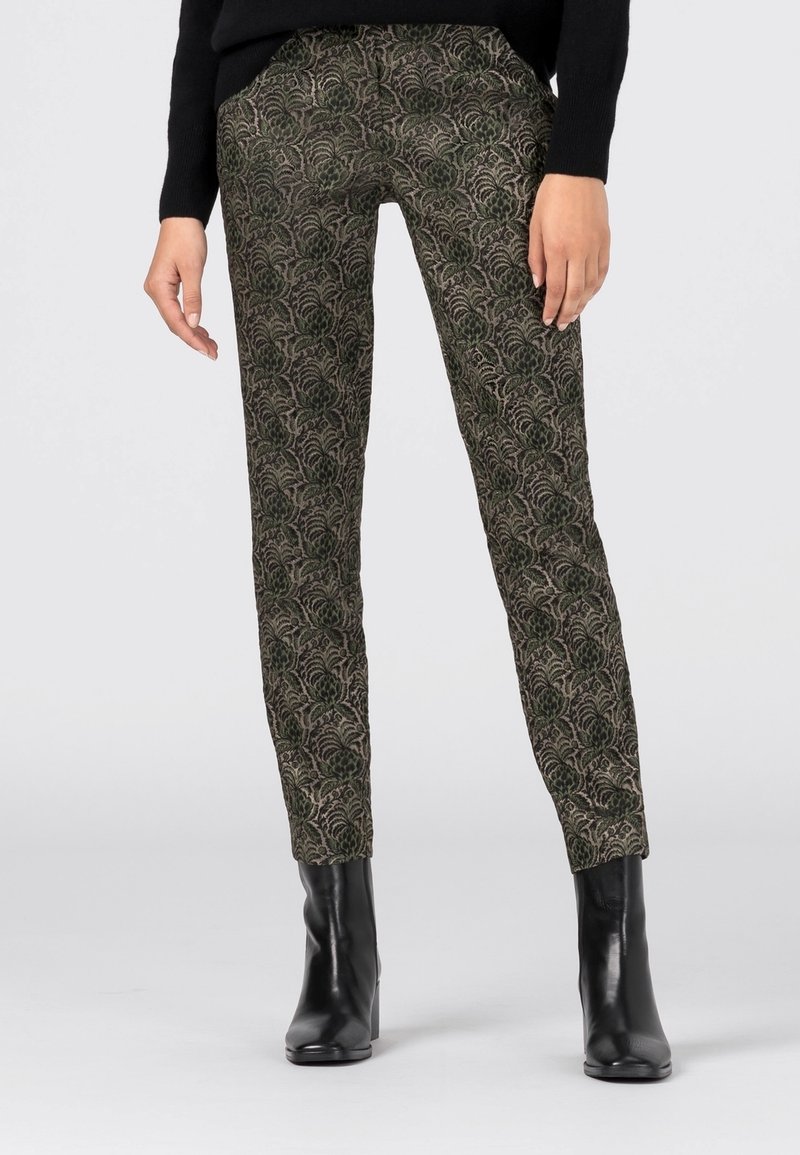 Femme portant un pantalon slim avec un motif complexe de feuilles vertes et noires, associé à des bottines noires à talons et un pull noir.