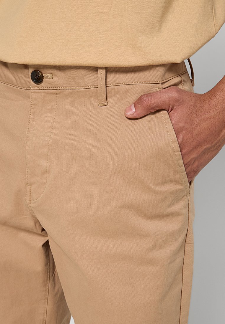 Khaki-Chinos aus glattem Baumwollstoff. Sie verfügen über einen einzelnen Knopfverschluss und Fronttaschen und zeichnen sich durch eine taillierte Passform und klare Linien aus.