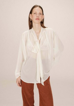 Blusa blanca holgada y transparente con un detalle de lazo en el cuello. Mangas largas con puños fruncidos. Combinada con pantalones de pana marrones.