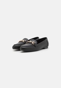 Mocassins en cuir noir avec une semelle plate, présentant un accent décoratif de chaîne dorée à l'avant et un design de bout arrondi et lisse.