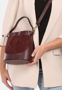 Sac à main bordeaux avec un corps en cuir texturé et un devant en daim doux. Il est doté d'une poignée et d'une sangle réglable, arborant une forme ronde.