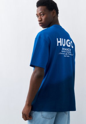 Νέος άνδρας που φοράει μπλε μπλουζάκι με το λογότυπο "HUGO" στην πλάτη, κοιτώντας πάνω από τον ώμο του σε απλό φωτεινό φόντο.