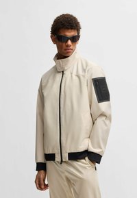 Giacca impermeabile beige con zip frontale, colletto alto, rifiniture nere e tasca con zip sulla manica sinistra. Materiale liscio e leggero.